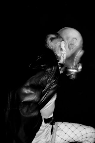 Person mit Gasmaske, Lederjacke und Netzstrümpfen vor schwarzem Hintergrund. Das Bild ist schwarz-weiß.