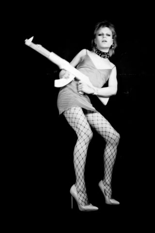 Eine Person in einem Kleid, Netzstrümpfen und High Heels posiert mit einer großen Spielzeugpistole vor einem schwarzen Hintergrund.