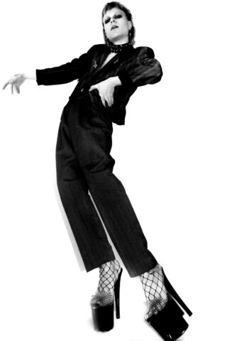 Person in schwarzer Jacke und Hose lehnt sich dramatisch nach hinten, trägt Netzstrümpfe und hohe Plateauschuhe, vor einem weißen Hintergrund.