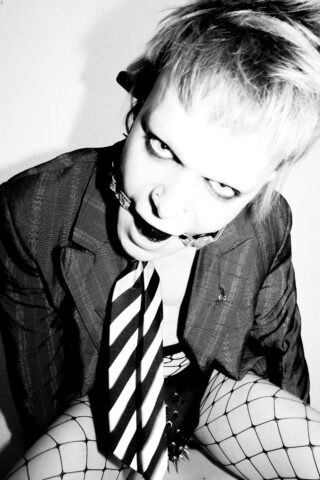 Person im Jackett, gestreifter Krawatte und Netzstrümpfen trägt einen Mundschutz und blickt in einem kontrastreichen Schwarz-Weiß-Foto mit eindringlichem Gesichtsausdruck in die Kamera.