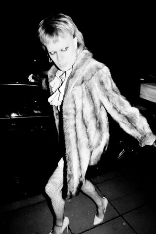 Eine Person in einem Kunstpelzmantel, Netzstrümpfen und High Heels geht nachts im Licht eines hellen Blitzes auf einem Bürgersteig.