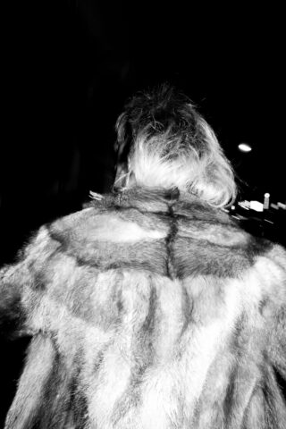 Eine Person mit hellem Haar ist von hinten zu sehen. Sie trägt einen gemusterten Pelzmantel und geht nachts spazieren. Das Bild ist schwarz-weiß und weist einen hohen Kontrast auf.