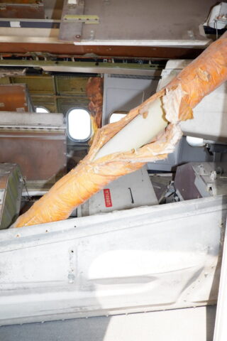 Im Inneren eines beschädigten Flugzeugs ist ein großer, orange isolierter Strukturträger sichtbar und zerbrochen, während in der Kabine zerrissene Isolierung und Schutt verstreut liegen.