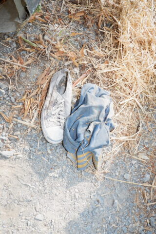 Ein abgetragener Turnschuh und ein zerknittertes blaues Hemd mit gelben Streifen liegen im Sonnenlicht auf Kies und trockenem Gras.