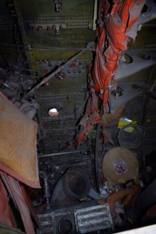 Innenraum eines alten, verlassenen Flugzeugs mit zerrissener orangefarbener Isolierung, freiliegenden Kabeln, Schutt und auf dem Boden verstreuten Warnschildern.