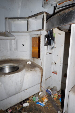 Schmutziges, überfülltes Inneres eines kleinen, heruntergekommenen Raums mit einem Waschbecken, Regalen und verstreutem Müll und Schutt auf dem Boden.