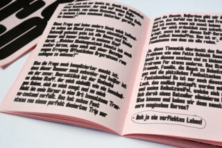 Ein aufgeschlagenes Heft mit deutschem Text in fetten, schwarzen Buchstaben auf rosa Seiten, aus einem leichten Winkel vor weißem Hintergrund fotografiert.