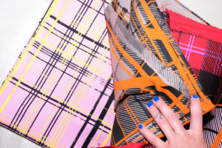 Eine Hand mit blau lackierten Nägeln hält zwei gemusterte Stoffstücke, eines mit rosa und gelbem Karomuster und das andere mit orange, schwarz und rot geometrischem Muster.