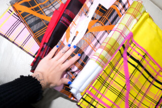 Eine Hand mit blauem Nagellack berührt mehrere bunte Stoffstücke mit Karomuster, die auf einer hellen Oberfläche ausgebreitet sind.
