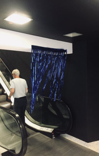Ein Mann steht neben einer Rolltreppe neben einem Türrahmen, der mit blauen, metallischen Fransenbändern bedeckt ist, die an der Wand darüber befestigt sind.