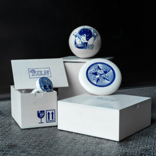 Drei weiße Schachteln und drei Bowlingkugeln mit blauen Grafiken und Text sind auf einem Metallboden vor dunklem Hintergrund angeordnet. Ein Schachteldeckel und eine Kugel sind geöffnet und aufrecht aufgestellt.