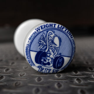 Ein weißes Keramikobjekt mit der blauen Illustration eines Bären, der eine 10 kg schwere Hantel hebt, die Worte „Weight Lifting“ und „FUN“ sind sichtbar, platziert auf einer strukturierten Metalloberfläche.