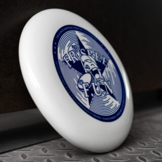 Ein weißer Frisbee mit einem blauen „Frisbee Fun“-Logo und einem Cartoon-Charakter-Design liegt aufrecht auf einer Metalloberfläche auf einem strukturierten Boden.