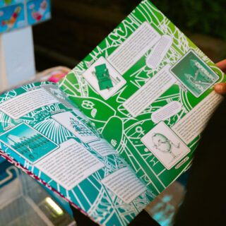 Eine Person hält ein buntes Buch mit grünen und weißen Seiten offen, das Text, Illustrationen und Diagramme enthält.