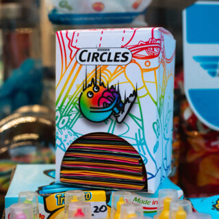 Zwischen anderen bunten Produkten befindet sich eine bunte Schachtel mit der Aufschrift „Hidden Circles“ mit einem Cartoon-Gesicht und einem Autogramm, die gestapelte runde Gegenstände enthält.