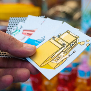 Eine Hand, die mehrere illustrierte Karten hält, wobei die oberste Karte ein gelb-weißes Schnellboot zeigt.