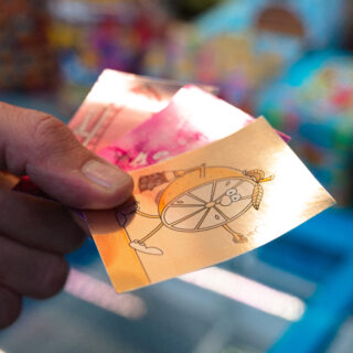 Eine Hand hält mehrere bunte Vergnügungs- oder Spielhallentickets, eines davon zeigt eine Zeichentrickfigur mit einem radähnlichen Körper.