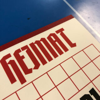 Eine Nahaufnahme eines Kalenders oder Planers mit dem Wort „HEIMAT“ in fetten, roten, eckigen Buchstaben auf weißem Hintergrund mit einer blauen Fläche darunter.