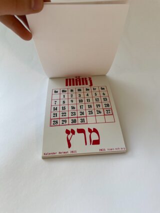 Eine Hand hebt eine Seite eines Kalenders für 2021 an, auf dem der Monat März auf Deutsch und Hebräisch angezeigt wird, wobei die Daten in einem Rasterformat dargestellt sind.