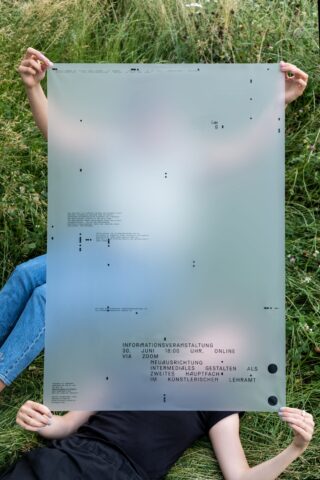 Zwei Personen liegen im Gras und halten ein großes durchsichtiges Laken mit aufgedrucktem Text und Symbolen in der Hand, das ihre Gesichter und Oberkörper teilweise verdeckt.