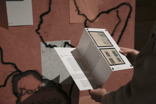 Eine Person hält eine offene Broschüre mit Bildern und Text vor einer Wand, an der eine Karte und Papiere angebracht sind.