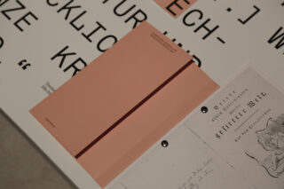 Auf einem Tisch liegt zwischen Papieren mit schwarz gedrucktem Text und einem handschriftlichen Dokument ein pfirsichfarbenes Heft mit einem dunklen Längsstreifen.