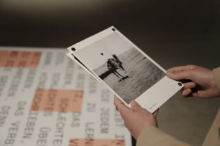 Eine Person hält ein Schwarzweißfoto einer Person auf einem Pferd, das über einem Tisch mit Papier steht, auf dem deutscher Text aufgedruckt ist.