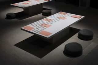 Niedrige Tische mit aufgedrucktem Text und orangefarbenen Plakaten sind in einem schwach beleuchteten Raum mit gefliestem Boden von runden schwarzen Bodenkissen umgeben.