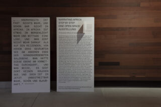 Zwei Informationstafeln mit deutschem und englischem Text stehen an einer Holzwand in einem schwach beleuchteten Raum.