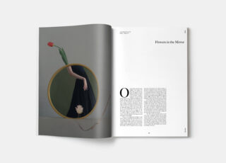 In einer aufgeschlagenen Zeitschrift ist auf der linken Seite ein Foto zu sehen, auf dem sich ein Frauenarm und eine Tulpe in einem runden Spiegel spiegeln, und auf der rechten Seite ein Artikel mit dem Titel „Blumen im Spiegel“.