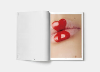 Aufgeschlagene Zeitschrift mit einer Nahaufnahme von Lippen, auf denen zwei glänzende, rote, herzförmige Objekte liegen; die linke Seite ist leer.
