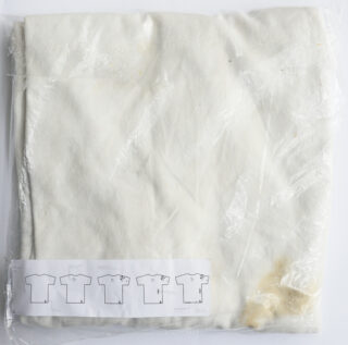 Ein gefaltetes, cremefarbenes Kleidungsstück in einer durchsichtigen Plastiktüte mit einem Papieretikett mit T-Shirt-Diagrammen und einigen sichtbaren gelben Flecken auf dem Stoff.