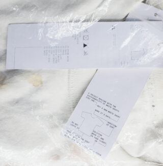 Weißes Hemd mit braunen Flecken, teilweise bedeckt von einer durchsichtigen Plastikverpackung und zwei rechteckigen Papieretiketten mit aufgedrucktem Text und Pflegehinweisen.