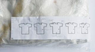 Ein in Plastik eingewickeltes weißes T-Shirt mit einem Etikett, auf dem fünf Strichzeichnungen von T-Shirts zu sehen sind, die jeweils unterschiedliche Flecken oder Verfärbungen sowie Textanmerkungen aufweisen.