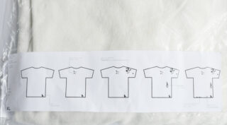Ein weißes Kleidungsstück in einer Plastikverpackung mit einem bedruckten Papier, auf dem fünf T-Shirt-Umrisse abgebildet sind, die jeweils mit unterschiedlichen Mustern oder Flecken versehen sind und in der unteren linken Ecke mit „XL“ beschriftet sind.