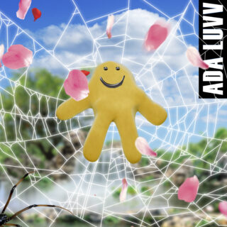 Eine gelbe, lächelnde Plüschfigur hängt im Zentrum eines Spinnennetzes im Freien, umgeben von herabfallenden rosa Blütenblättern. Am rechten Rand steht der Name „ADA LUVV“.