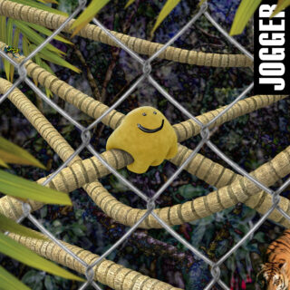 Eine gelbe, blobartige Kreatur mit einem Smiley-Gesicht sitzt auf dicken Ranken hinter einem Maschendrahtzaun in einer Dschungelkulisse; am rechten Rand erscheint das Wort „JOGGER“.