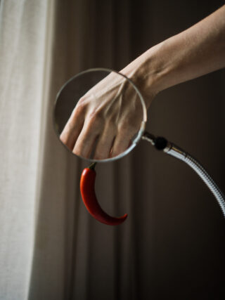 Durch eine Lupe wird eine Hand betrachtet, am Griff der Lupe hängt eine rote Chilischote.