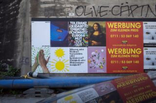 Eine Wand mit verschiedenen Plakaten, darunter Anzeigen, Kunstwerke und Texte auf Deutsch; darüber sind Graffiti und im Vordergrund sind blaue Rohre und ein Autodach zu sehen.
