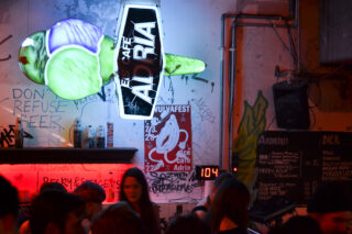 Über einem überfüllten Barbereich mit Graffiti und Postern an der Wand hängt ein Neonschild mit der Aufschrift „EIS CAFE ADRIA“.