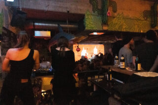 Eine schwach beleuchtete Bar mit geselligen Gesprächen, einem DJ, der Musik spielt, einer Menschenmenge, die vor einer hell erleuchteten Bühne steht, und auf der Theke stehen Bierflaschen.