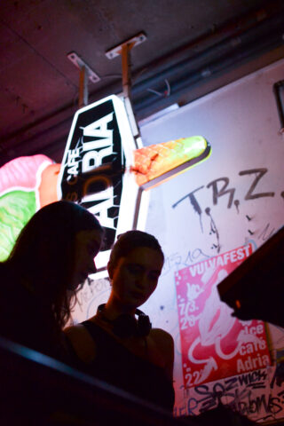 Zwei Personen stehen in der Nähe von DJ-Equipment in einem schwach beleuchteten Raum mit Graffiti-bedeckten Wänden und einem Neonschild „Adria“ im Hintergrund.