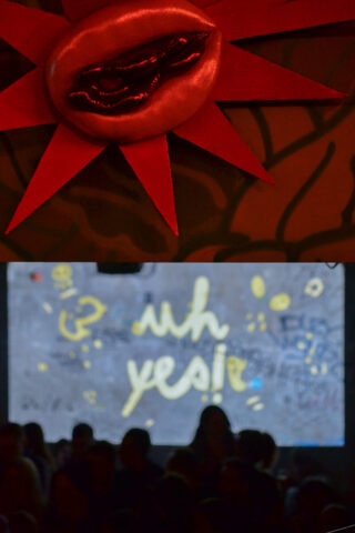 Oben ist ein dekoratives rotes Lippenornament mit Strahlen zu sehen, darunter zeigt ein Bildschirm in gelber Schrift „Oh ja!“ an, davor sind Silhouetten von Menschen zu sehen.