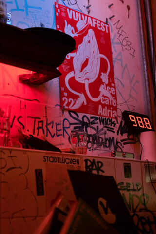 Ein rotes Plakat für das „Vuvafest“ hängt an einer mit Graffiti bedeckten Wand über einem Regal mit einer Digitaluhr mit der Anzeige 98,8, Büchern und verschiedenen Aufklebern. Die Szene ist mit rotem Licht beleuchtet.