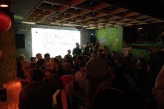 Eine Menschenmenge versammelt sich in einer schwach beleuchteten Bar oder einem Club mit Graffiti an den Wänden und einem großen, beleuchteten Herz im Hintergrund.