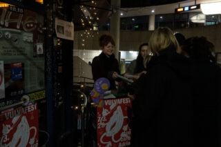 Eine Gruppe von Menschen steht nachts in der Nähe eines mit Plakaten bedeckten Türrahmens; eine Person verteilt Gegenstände, während sich andere unter Lichterketten darum versammeln.