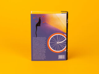 Ein Buch mit violettem Einband, auf dem vor einem einfarbig gelben Hintergrund eine schwarze Katze mit orangefarbenen Augen und einem Fahrradrad abgebildet ist.