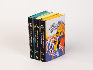Drei gebundene Bücher stehen aufrecht; zwei haben einen schwarzen Buchrücken mit weißem Text und eines hat einen bunt illustrierten Einband mit zwei Personen auf einem Fahrrad.