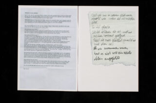 Ein aufgeschlagenes Heft zeigt auf der linken Seite einen getippten Brief und auf der rechten Seite eine handschriftliche Notiz in deutscher Sprache auf einem zerrissenen Stück Papier.