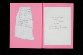 Ein offenes rosa Buch mit handgeschriebenem deutschen Text auf zwei Seiten, eine auf einem abgerissenen Stück Papier und die andere direkt auf der Seite.